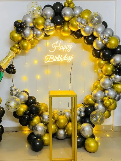 A fun champagne-themed birthday party decoration. The setup includes a balloon ring in black, gold, and silver, with a large champagne bottle foil balloon.