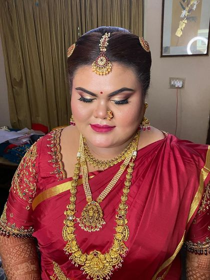 A close-up of a bride's muhurtham look, showing the neat updo that provides a secure base for the maang tikka and head jewellery. The red silk saree is draped to complement the heavy gold necklaces.
