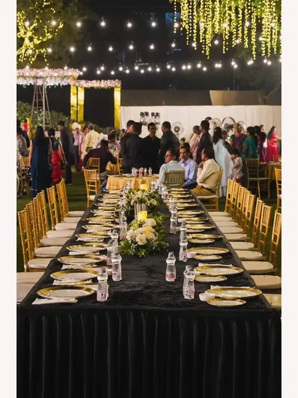 An intimate long-table setup for a special sit-down dinner under the stars. The black tablecloth, golden plates, and ambient lighting create a sophisticated and memorable dining experience.