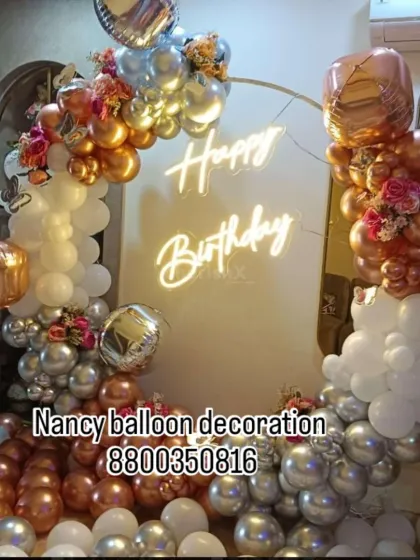 A glamorous ring backdrop using chrome balloons in rose gold and silver. The arrangement is accented with artificial flowers and a 'Happy Birthday' neon sign for a touch of luxury.