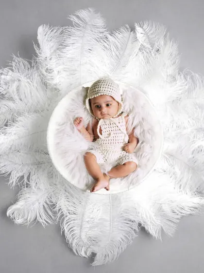 A creative overhead shot of a baby in a white knitted outfit, surrounded by a circle of soft white feathers. This creates a beautiful, angelic look.