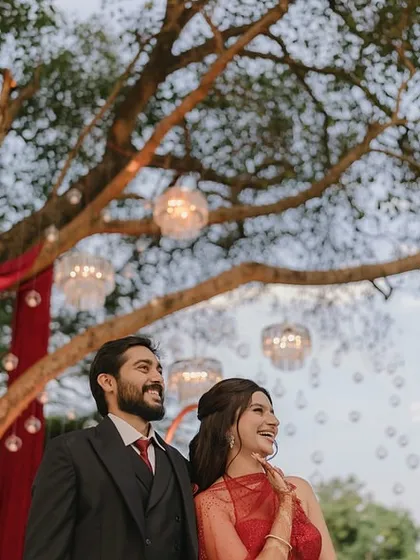A happy couple posing under a tree adorned with hanging glass bubbles and red drapes at their engagement party.