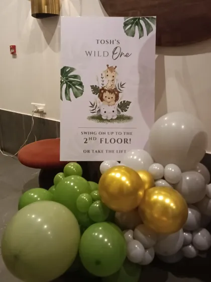 A charming welcome sign for a "Wild One" jungle-themed party. The sign directs guests with a cute message, complemented by a balloon cluster in theme-appropriate green, white, and gold.