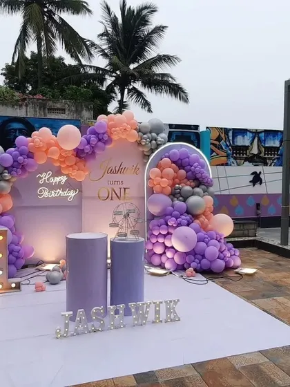 An outdoor first birthday setup in a lovely lavender and peach color scheme. The decor includes a neon "Happy Birthday" sign, a light-up number "1", and personalized name blocks.