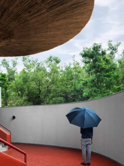 A solitary figure with an umbrella stands within the curved corridor of the TAPMI Centre. The red oxide floor contrasts with the grey walls and lush green view, creating a rich sensory experience.