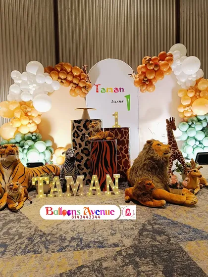 A safari-themed first birthday for Taman in a party hall. The setup includes animal print cake stands, a variety of large animal props, and a balloon arch in earthy tones.