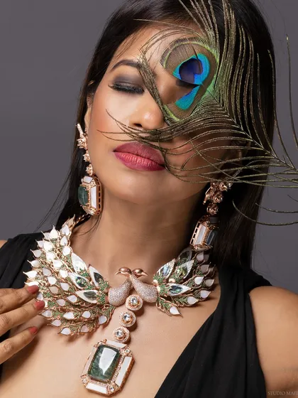 A creative beauty shot focusing on an elaborate peacock-themed statement necklace, with a real peacock feather artfully placed over the model's eye. This image is perfect for showcasing high-end jewellery and accessory campaigns.