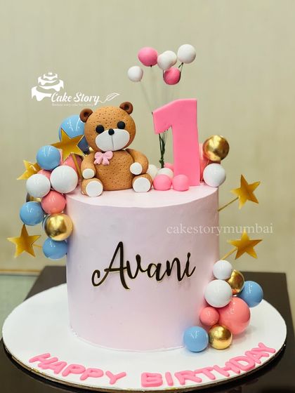 A sweet teddy bear cake for Avani's first birthday. The pink cake is topped with a fondant teddy bear and decorated with colorful balloons in blue, pink, and gold.