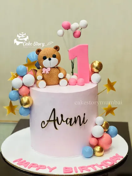 An adorable teddy bear cake for a first birthday. This tall pink cake is decorated with a fondant bear and colorful balloon clusters in pink, blue, and gold.