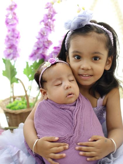 A close-up shot capturing the sweet connection between two sisters. The older sister proudly holds her new baby sibling, who is wrapped in a lovely lavender swaddle.