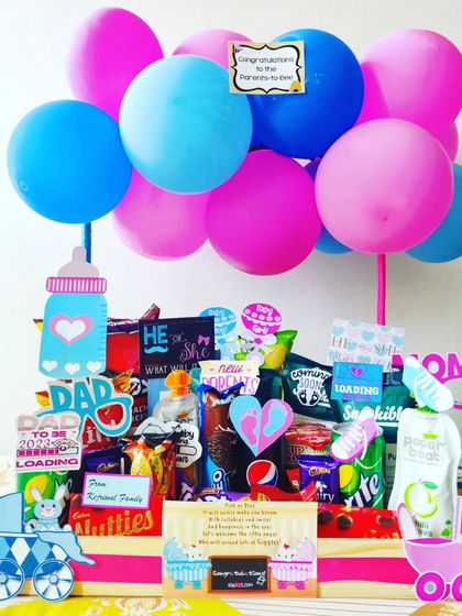 This large hamper is a feast for the parents-to-be, loaded with drinks, snacks, and chocolates. The pink and blue balloons and baby-themed cutouts make it a festive centerpiece for a baby shower.