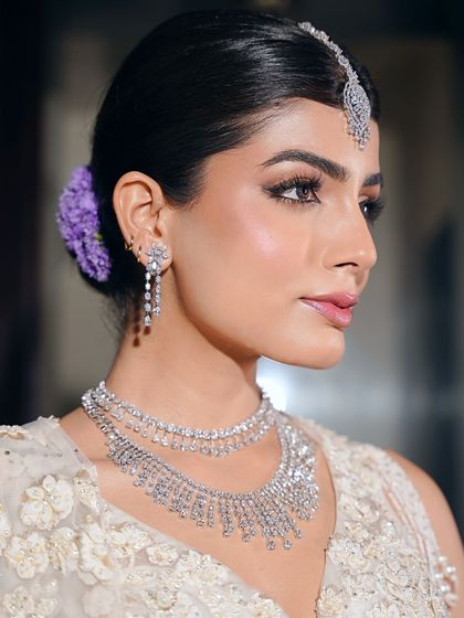 A close-up of the dewy bridal glam. The skin is luminous, the eyes are softly defined, and a hint of purple flowers in her hair adds a unique, modern touch to the classic white bridal look.