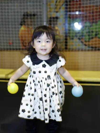 A beautifully dressed toddler enjoys a moment of fun on our trampoline, holding two colorful balls.