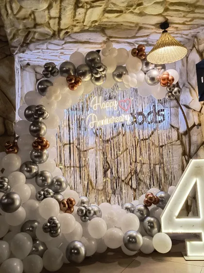 A rustic stone wall provides a unique background for this white, silver, and chrome balloon arch. The setup includes a shimmer curtain, a neon sign, and a large marquee number, all of which can be tailored for your anniversary theme.