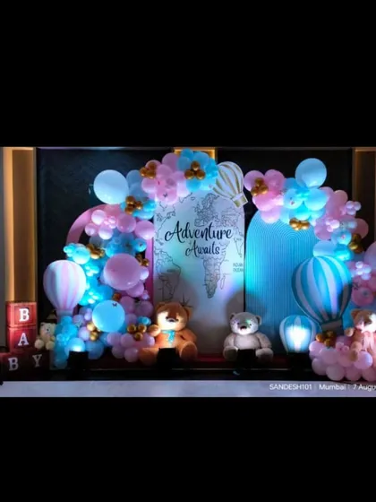 An "Adventure Awaits" baby shower theme. This setup features a world map backdrop, hot air balloon props, and a pink and blue balloon arch.