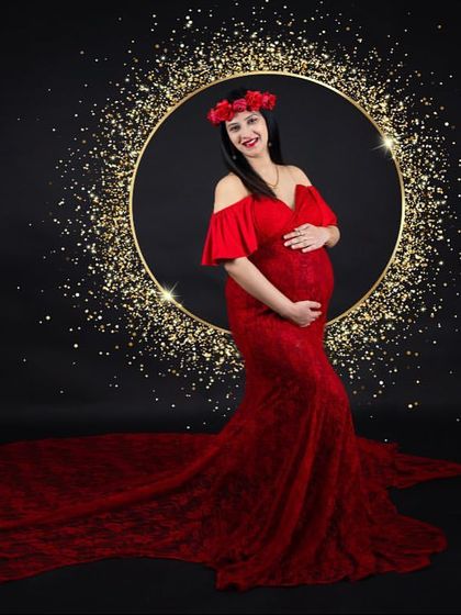 A dramatic and glamorous shot. The mom-to-be stands in a red lace gown with a floral crown, framed by a golden ring of sparkling lights against a black background.