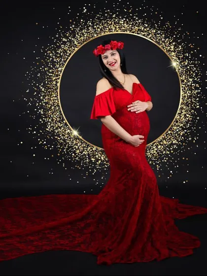 A dramatic and glamorous shot. The mom-to-be stands in a red lace gown with a floral crown, framed by a golden ring of sparkling lights against a black background.