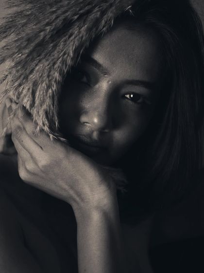 A moody and mysterious black and white portrait. The pampas grass partially obscures the model's face, and the deep shadows create an intimate, secretive atmosphere. The focus is on the eye that peeks through.