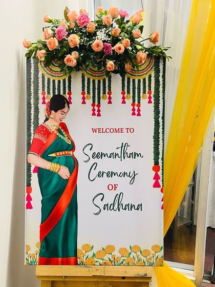 A traditional welcome board for a Seemantham ceremony. The design features an illustration of a mother-to-be in a saree, with marigold and floral garland motifs.