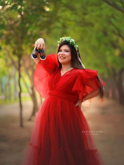 A fun and sweet outdoor shot, with the mother-to-be holding up a pair of baby shoes. The red gown and floral crown add to the charm.