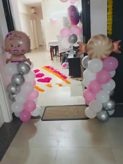A sweet entrance setup for a baby girl's arrival. Balloon pillars with baby and candy foil balloons stand on either side of a flower petal rangoli, creating a cheerful welcome.