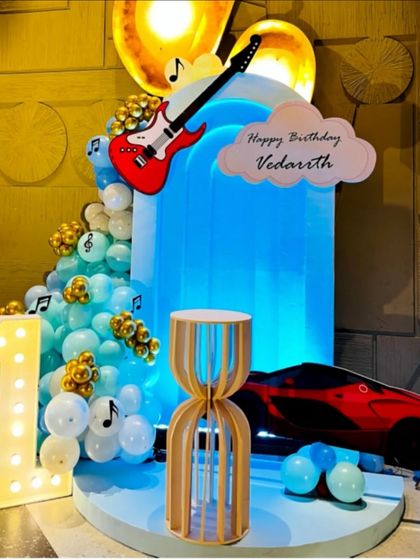 A unique theme combining music and motors for Vedarath's first birthday. The stage features a red sports car, a guitar cutout, musical notes, and cool blue lighting for a stylish celebration.