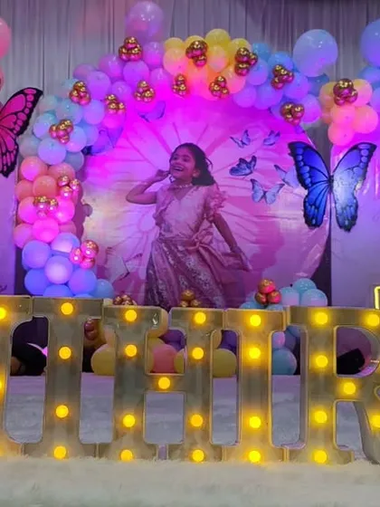 Let's make your child's birthday magical with a butterfly theme. This setup features a colorful balloon arch, a custom backdrop with the birthday girl's photo, and her name 'MIHIRA' spelled out in large, bright marquee letters for a personalized centerpiece.
