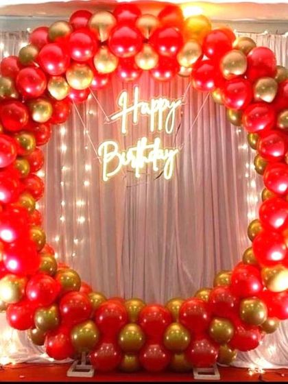 A classic red and gold balloon ring backdrop for a birthday party. Set against a curtain with fairy lights, this full and vibrant arrangement creates a bold and festive statement.