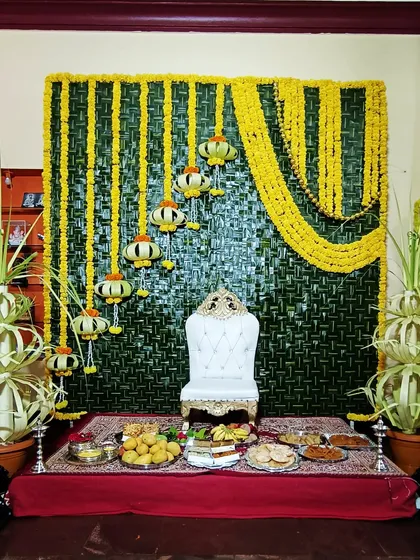 This traditional baby shower setup features a backdrop woven from fresh coconut leaves. The decor is completed with yellow floral arrangements, a special white throne for the mom-to-be, and a table of offerings.