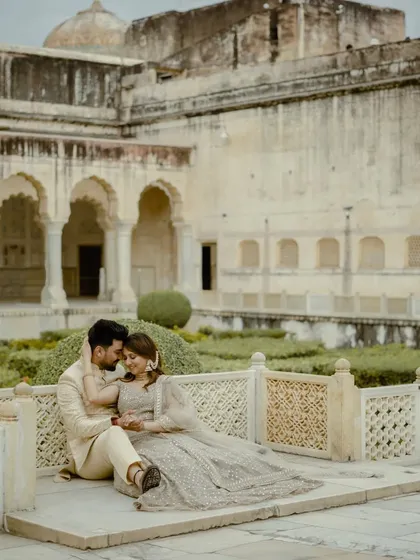 A serene and romantic portrait of the couple seated in a palace courtyard. The lush green gardens and ancient architecture provide a peaceful and beautiful setting.