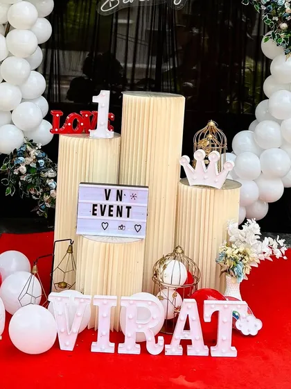 A close-up of a first birthday decoration for 'VIRAT'. This setup includes ribbed stands, a custom 'VN Event' lightbox, and other small props like a crown and 'LOVE' sign.