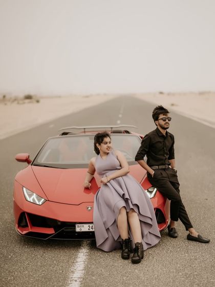 A wide shot of the couple with the Lamborghini on an empty desert road, emphasizing the sense of adventure.