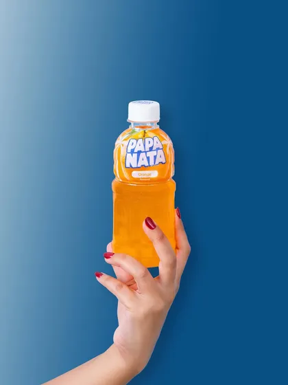 A clean, minimalist shot of a hand holding a Papa Nata orange drink against a solid blue background. This high-contrast, graphic style is modern and scroll-stopping.