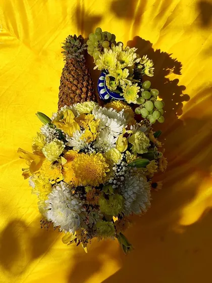 A creative flat lay for a Haldi theme, using a pineapple, grapes, and a bouquet of yellow and white flowers against a bright yellow fabric.