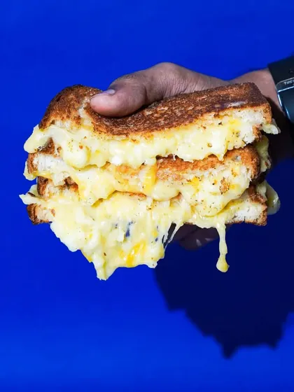 A hand holding a thick, cheesy grilled cheese sandwich against a bold blue background. The direct flash and vibrant color make this a simple but powerful, eye-catching shot.