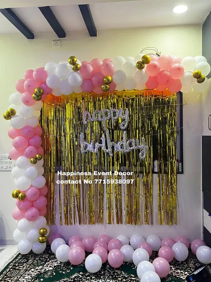An L-shaped balloon arch over a foil curtain is a classic party decoration. This setup uses pink, white, and gold balloons to create a simple and beautiful backdrop for a birthday celebration at home.