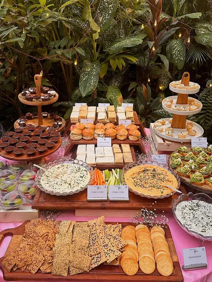 A beautiful outdoor setup for a garden party. The pink tablecloth and lush greenery provide a perfect backdrop for our assortment of dips, crackers, sandwiches, and desserts.