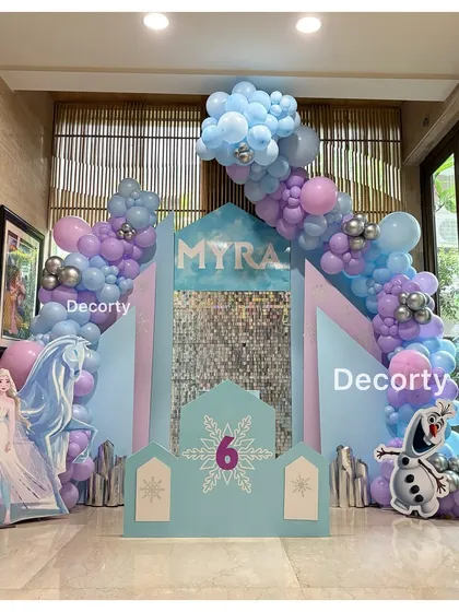 A winter wonderland for Myra's 6th birthday with this Frozen theme. The backdrop features an ice castle with a shimmering sequin panel, surrounded by blue, purple, and silver balloons.