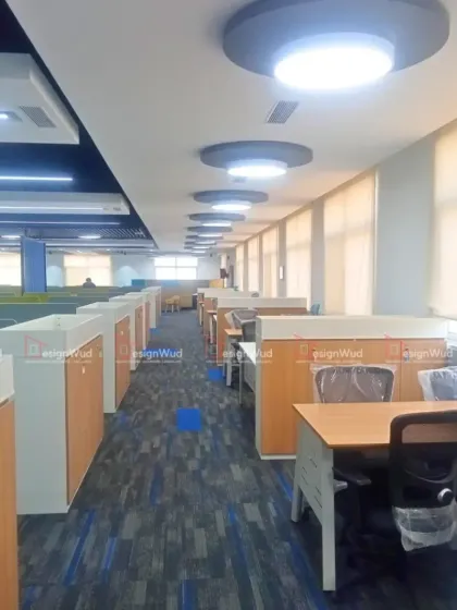 This perspective shows the organized layout of workstations along a windowed wall, maximizing natural light for employees. The ceiling features modern, circular LED light fixtures that provide even, glare-free illumination throughout the workspace.