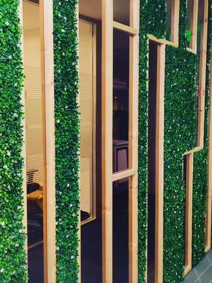 A custom office partition wall combining wood frames with vertical strips of artificial greenery. This design allows light to pass through while still providing a sense of separation.