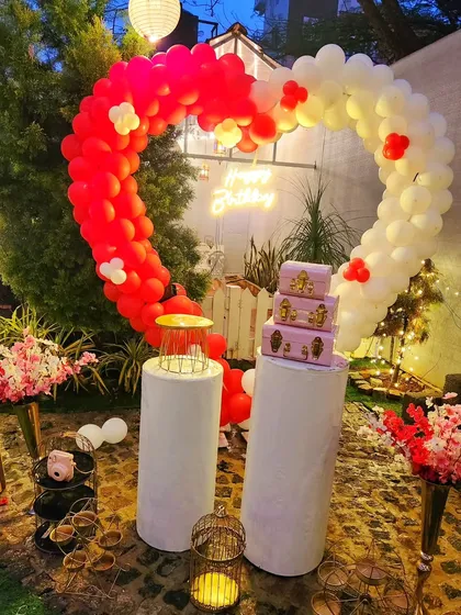 A vibrant birthday backdrop featuring a heart-shaped balloon arch in red and white. The setup includes pedestals for gifts or a cake and is accented with cherry blossom floral arrangements.