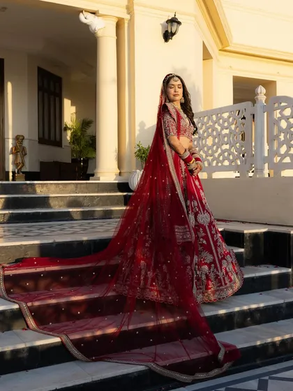A classic bridal portrait showcasing the bride, Sushmita, in her magnificent red lehenga. The long, flowing veil and regal pose against the palace architecture create a timeless and powerful image of bridal elegance.