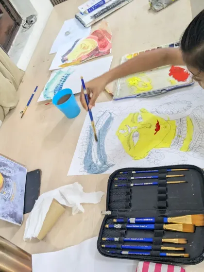 A top-down view of the creative process. The student is using a large brush to apply a blue wash, with her full set of brushes and palette ready for action.