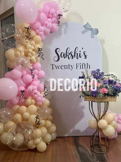 A beautiful pastel-themed setup for a 25th birthday. This decoration features a custom arched backdrop personalized with the name 'Sakshi', framed by a delicate balloon arrangement in shades of pink and nude.