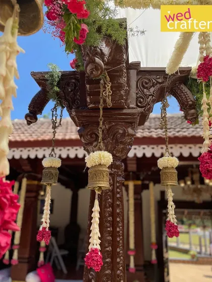 A detailed shot of a carved antique wooden pillar from the mandap, adorned with traditional flowers and brass bells, showcasing the craftsmanship.