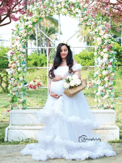 A beautiful solo portrait in front of a floral arch. The mother-to-be is wearing a white ruffled gown and holding a basket of flowers.