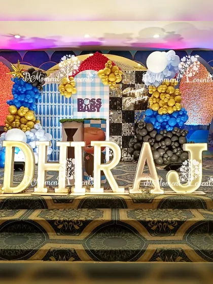 A dynamic Boss Baby themed stage with a checkerboard and gingham backdrop. The setup is brought to life with colorful balloon clusters and large, illuminated marquee letters.