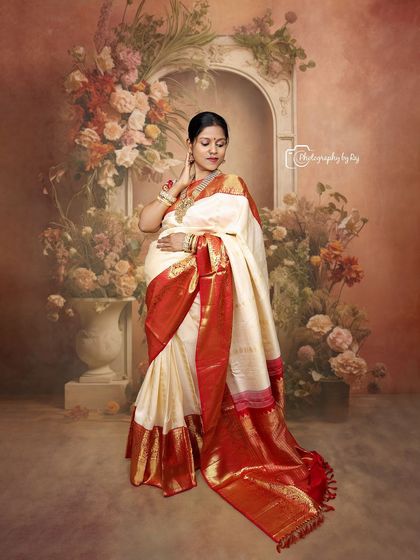 A beautiful studio portrait of a mother-to-be in a traditional red and white saree, against a floral painted backdrop.