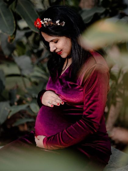 A quiet, reflective moment captured through foliage. The mother-to-be gently holds her belly, creating a very personal and intimate portrait.