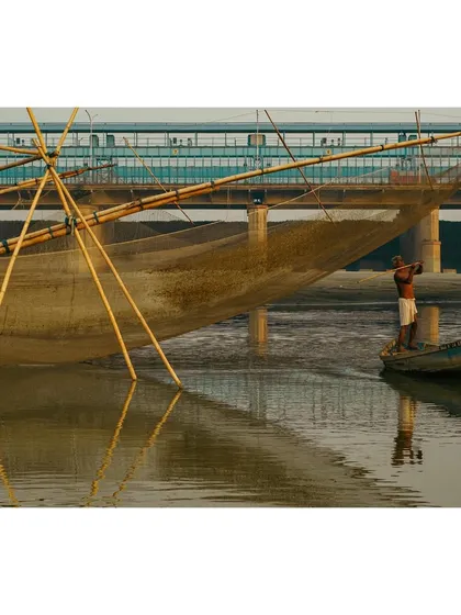 A fisherman tends to his large fishing net near Kalindi Kunj, with the metro bridge visible in the background. The reflection of the net in the water creates a beautiful symmetry.
